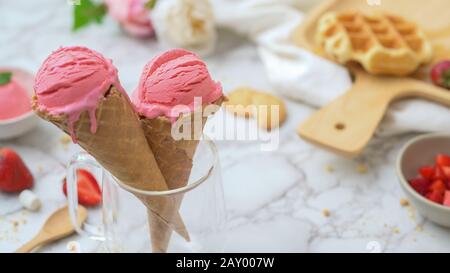 Vista ravvicinata del dessert estivo con coni gelato alla fragola, waffle e farcitura sulla scrivania in marmo Foto Stock
