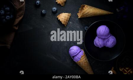 Vista dall'alto del dessert estivo con gelato al mirtillo, farcitura, cono di waffle e spazio di copia su sfondo nero da scrivania Foto Stock