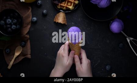 Vista dall'alto di una ragazza che tiene il cono gelato mirtillo con mirtillo, waffle e farcitura su sfondo nero tabella Foto Stock