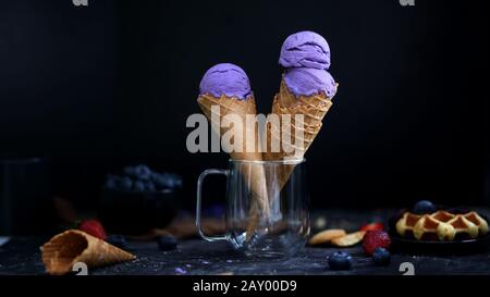 Vista ravvicinata del dessert estivo con coni gelato al mirtillo, waffle e farcitura su sfondo nero da scrivania Foto Stock