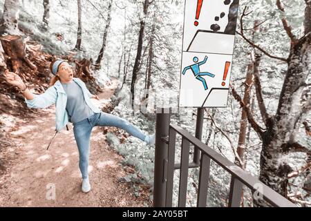 Divertente ragazza scivolata sul pendio scivoloso di un sentiero escursionistico in montagna vicino al segno. Concetto di rischio e sicurezza Foto Stock