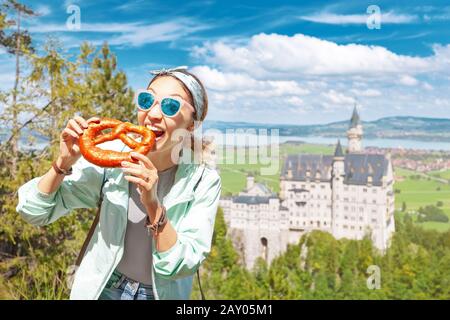 La ragazza felice del viaggiatore asiatico mangia prettzel croccante sullo sfondo del leggendario castello di Neuschwanstein. Turismo in Baviera concetto Foto Stock