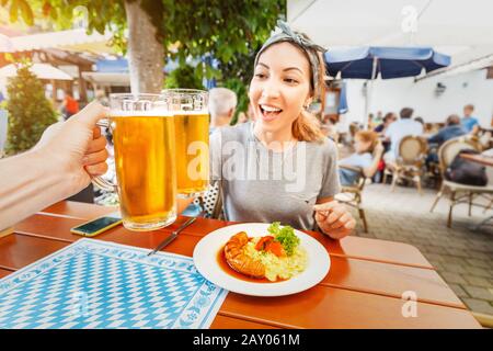 La ragazza asiatica felice ha graffato una tazza di birra bavarese in Biergarten. Tradizionale spuntino tedesco con salsiccia e insalata di patate Foto Stock