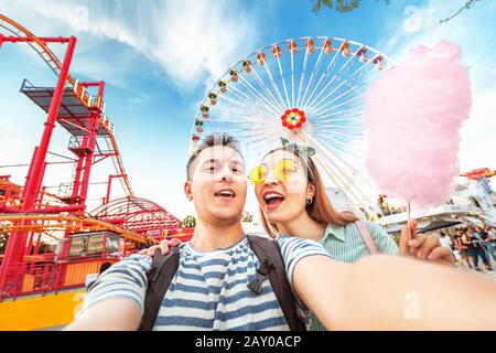 Amorevole coppia di razza mista abbracciando e mangiando caramelle di cotone e prendendo selfie durante il fine settimana al parco divertimenti. Data e relazione Foto Stock