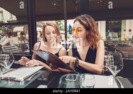 Due belle ragazze in abiti da sera seduti al ristorante e lettura menu insieme Foto Stock