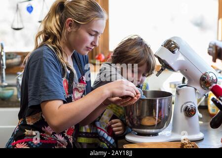 La ragazza teenage di tredici anni ed il suo fratello di 6 anni nella cucina, usando una ciotola di miscelazione Foto Stock