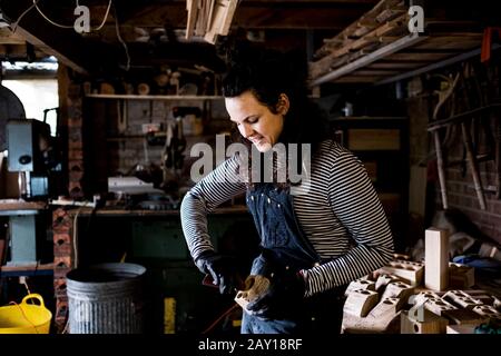 Donna con lunghi capelli marroni con ungaree, che lavora in laboratorio di legno. Foto Stock