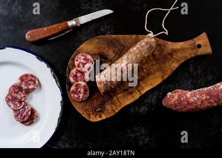 Alto angolo di chiusura del coltello, salame affettato su tagliere in legno e piastra in smalto bianco su sfondo nero. Foto Stock