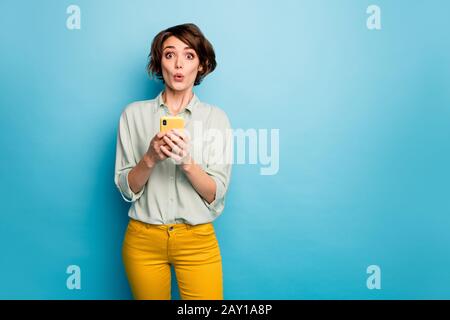 Foto di signora senza parole tenere le mani del telefono leggere nuovo blog commenti non credo occhi terrorizzati indossare casual verde camicia pantaloni giallo isolato blu Foto Stock