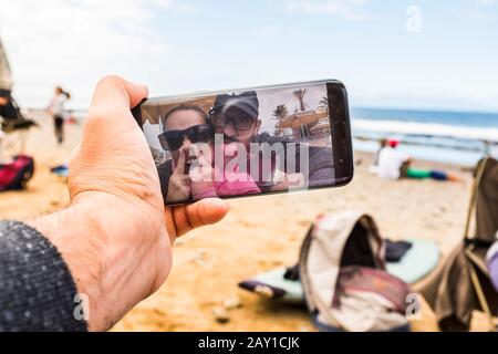 Il punto di vista dell'uomo di una divertente e folle coppia divertirsi prendendo selfie o fare video conferenza chiamata dalla spiaggia utilizzando una tecnologia moderna collegato sma Foto Stock