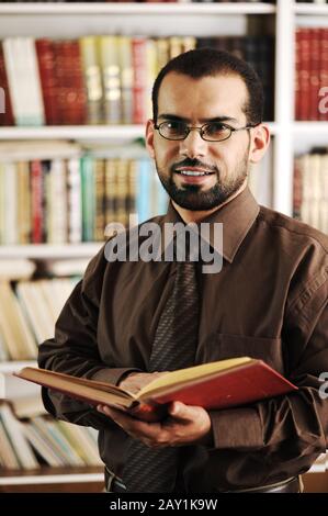 Giovane felice uomo in piedi nella biblioteca universitaria lettura e sorridente Foto Stock