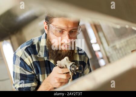 Falegname come pittore che usa la pistola di vernice mentre dipinge il legno nel negozio di vernice Foto Stock