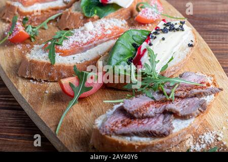 Panini con pesce rosso, manzo in suvit e formaggio morbido. Guarnita con fette di verde, rucola e pomodori. Su un vassoio ovale in legno. Copiare lo spazio Foto Stock