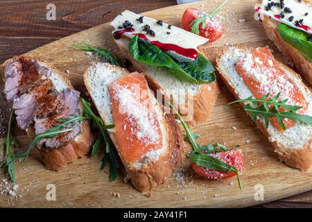 Panini con pesce rosso, manzo in suvit e formaggio morbido. Guarnita con fette di verde, rucola e pomodori. Su un vassoio ovale in legno. Copiare lo spazio Foto Stock