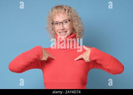 Donna sorridente divertente in occhiali che punta le dita su se stessa isolato su uno sfondo blu Foto Stock