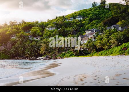 Mahe, Seychelles. Esotica spiaggia tropicale Anse Intensance al tramonto in serata luce. Vista panoramica degli alloggi di lusso nascosti nella giungla Foto Stock