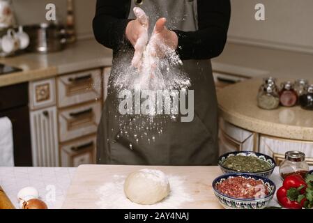 Una donna prepara l'impasto a casa e le stringe le mani con la farina. La farina scattera la cucina della casalinga. Foto Stock
