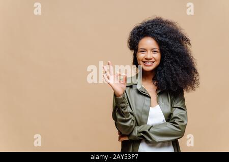 All'interno scatto di donna curly aspetto piacevole ha sorriso piacevole, fa il gesto giusto, segno eccellente, dà approvazione, vestito in shi di pelle alla moda Foto Stock