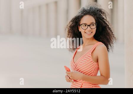 Felice millennial ragazza con capelli ricci, indossa abito rosa, occhiali ottici, messa a fuoco a parte con sorriso, stand all'aperto, aspetta per la chiamata, gode di alta velocità Foto Stock