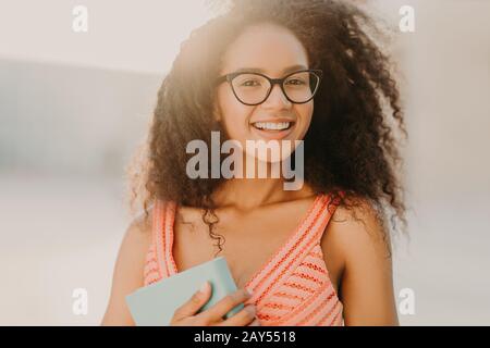 Foto di spensierata studentessa afroamericana in abito estivo, tiene libro, si prepara per l'esame, passeggi per le strade della città, gode di una calda giornata di sole, ha cha Foto Stock