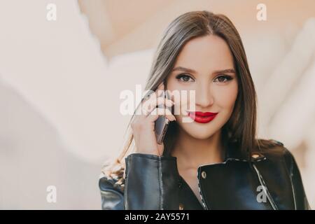 Primo piano scatto di bella donna parla con l'amico tramite app mobile, ha trucco e labbra rosse, vestito in pelle giacca, ha tempo libero all'aperto. Com online Foto Stock