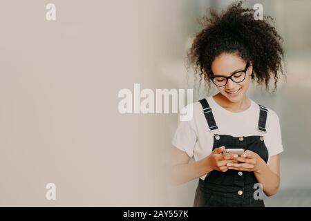 Bella ragazza afroamericana in abbigliamento alla moda, utilizza il telefono cellulare per inviare messaggi di testo, collegato al wi-fi, casella di controllo e-mail, si trova vicino Foto Stock