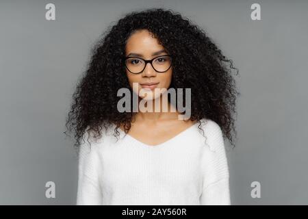 Ritratto di donna seria con pelle scura, capelli afro bush, indossa grandi occhiali trasparenti e maglione bianco morbido, guarda direttamente alla fotocamera, isolato ov Foto Stock