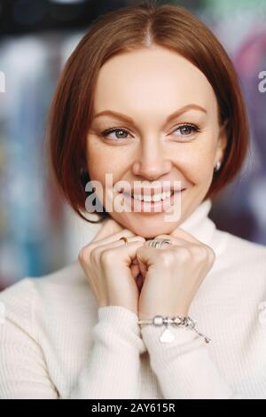 Foto di attraente giovane femmina con capelli castani diritti, guarda felicemente lontano, vestito con abiti bianchi, indossa il maglione dolcevita bianco, mantiene le mani u Foto Stock