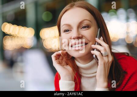 Headshot di attraente giovane donna con felice espressione facciale, parla via telefono cellulare, ha capelli marroni, felice di sentire le notizie, ha tempo libero, backg sfocato Foto Stock