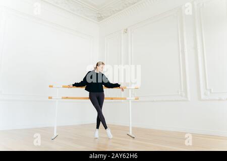 Ballerino classico ha stretching workout, stand vicino al balletto barres, indossa felpa nera e leggings, fa esercizi in studio, messo a fuoco giù. Foto Stock