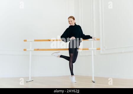 Persone, danza, coreografia, concetto di flessibilità. SLIM felice rosso capelli femmina in costume nero, solleva una gamba, si erge vicino al balletto barre in emp Foto Stock