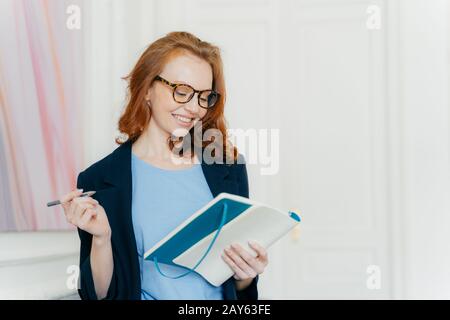 Felice zenzero giovani piani di lavoro, scrive in taccuino, fa note di informazioni utili, tiene la penna, ha sorriso sul viso, vestito elegante Foto Stock