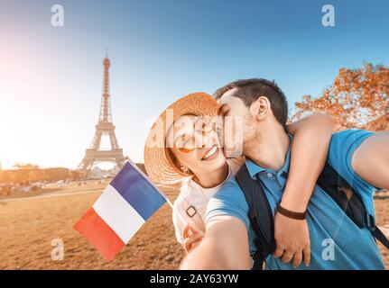 Coppia mista di gara in amore abbracciando e prendendo selfie con la bandiera francese e la Torre Eiffel sullo sfondo. Viaggio di nozze in Francia e Parigi Concept Foto Stock
