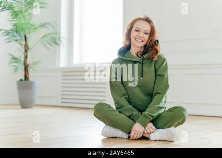 Un tocco orizzontale di allegra sportswoman in tuta verde, sneakers bianche, si siede in posa lotus, ha un sorriso amichevole sul viso, posa al coperto. Femmina Foto Stock