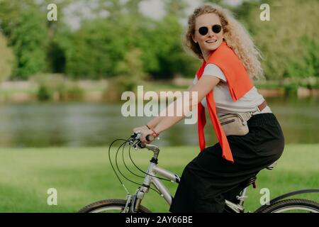La donna curly caucasica attiva guarda da parte, corre in bicicletta sportiva, copre la distanza e viaggia all'aperto, vestita con abiti casual, pone vicino a piccoli ri Foto Stock