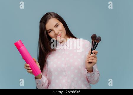 Felice giovane modello femminile fa acconciatura e trucco, contiene bottiglia di spazzola per capelli e cosmetici, testa tilts, vestito con abiti eleganti, posa o Foto Stock