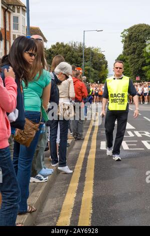 Corridori e spettatori che si raccolgono in strada pochi minuti prima dell'inizio di UNA corsa di beneficenza 10KM e il direttore di gara indossa una giacca ad alta visibilità. Foto Stock