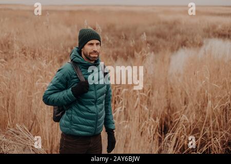 Colpo all'aperto di un viaggiatore maschio pensivo vestito con abiti caldi, di buon umore, ha zaino sul retro, passeggiate in ambiente rurale vicino campo durante l'autu Foto Stock