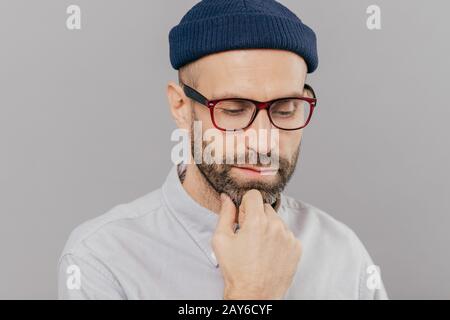 Colpo di Testa di sad lonely grave maschio mantiene la mano sotto il mento, ha setole scure, guarda giù, pensa di promozione e di avviamento nuova carriera, indossa spectac Foto Stock