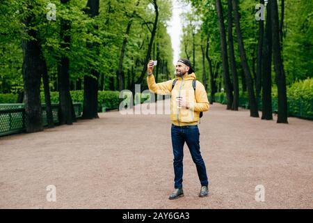 Ritratto a tutta lunghezza di giovane bearded vestito in giacca gialla, jeans e cappuccio nero dopo aver camminato attraverso il parco verde facendo selfie con il suo smartphon Foto Stock