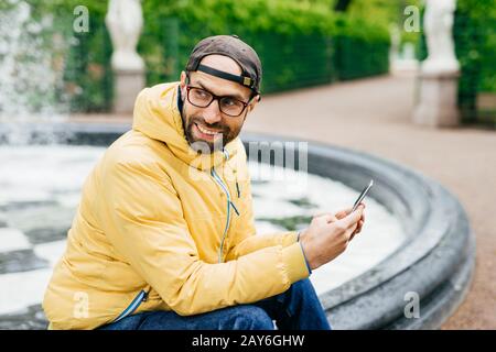 Ritratto laterale esterno di allegro uomo elegante bearded in giacca gialla e occhiali seduti vicino alla fontana nel parco tenendo moderna decisione nelle mani Foto Stock
