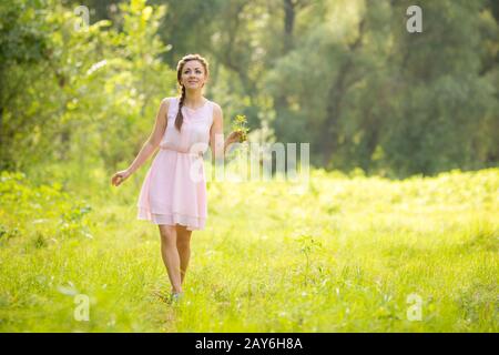 Giovane bella donna che cammina su un prato verde con un mazzo di fiori selvatici in una luce abiti estivi Foto Stock