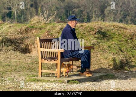 un anziano gentiluomo anziano seduto su una panchina di legno al mare con il suo cane seduto sotto la panca su un piombo. Foto Stock