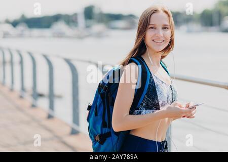 Sorridente bella donna con espressione felice, indossa casual top, porta borsa, tiene il telefono cellulare in mano, gode di comunicazione online con gli amici, bei Foto Stock