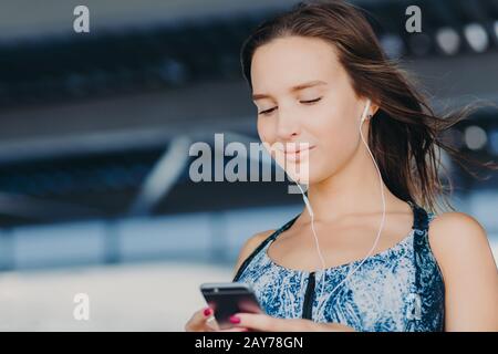 Primo piano ritratto di attraente giovane donna tiene smartphone, testi feedback, controlli casella e-mail, ascoltare musica nel lettore, download news canzoni, connessione Foto Stock