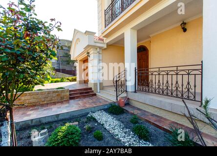 La facciata di un cottage classico bianco a due piani con giardino frontale. Porta d'ingresso scolpita, cancello auto, piastrelle beige, alberi. Foto Stock