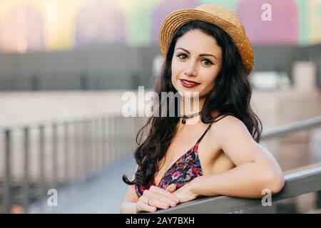 Bella donna con capelli lunghi scuri, aspetto accattivante indossando cappello estivo e vestito con escursione in grande città che posa al ponte. Giovani donne in viaggio Foto Stock