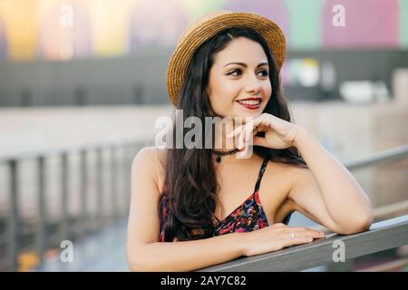 Giovane modello femminile con lunghi capelli scuri spessi in piedi al ponte con espressione sognante guardando in lontananza, indossando cappello estivo e abito a riposo o Foto Stock