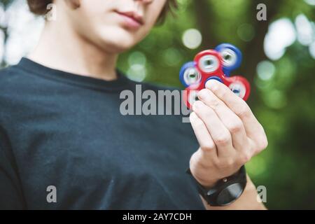 Un hipster dai capelli lunghi in un cappuccio si siede su una panchina e fa girare un fidget-spinner Foto Stock