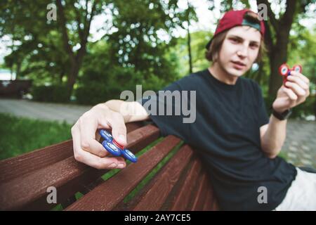 Un hipster dai capelli lunghi in un cappuccio si siede su una panchina e fa girare un fidget-spinner Foto Stock